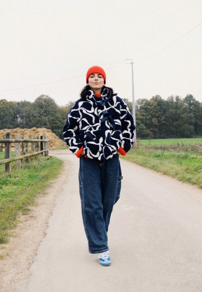 Abrigo de lana borroso en color navy con patrones abstractos blancos, gorro rojo, camiseta roja, pantalones de pana azul holgados y zapatillas azules, de pie en un camino de tierra.