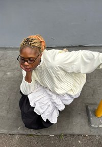 Top branco plissado sobreposto a uma saia branca e preta com folhos. Pessoa agachada com o queixo na mão, usando óculos e um acessório de cabelo.