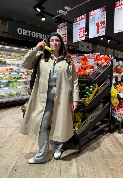 Abrigo trench beige llevado sobre una sudadera gris y pantalones de chándal acampanados. De pie en un mercado, sosteniendo un plátano junto a los expositores de frutas.