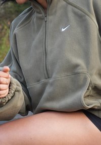 Personne portant une veste polaire Nike vert olive avec une fermeture éclair à l'avant, assise avec un genou visible, en extérieur avec un arrière-plan de verdure flou.