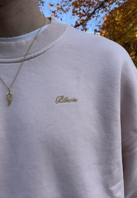Lichtroze sweatshirt met geribde halslijn en goud geborduurd logo. Heeft een gladde textuur, afgebeeld tegen een vervaagde natuurlijke achtergrond.