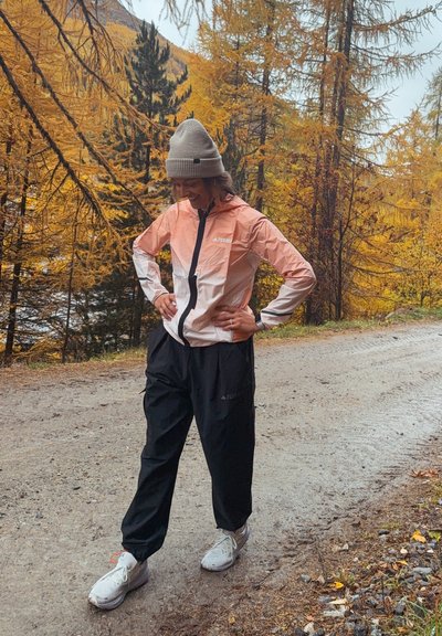 Chaqueta ligera y resistente al agua en rosa y blanco, combinada con pantalones negros. Presenta capucha, cierre de cremallera y acentos reflectantes.
