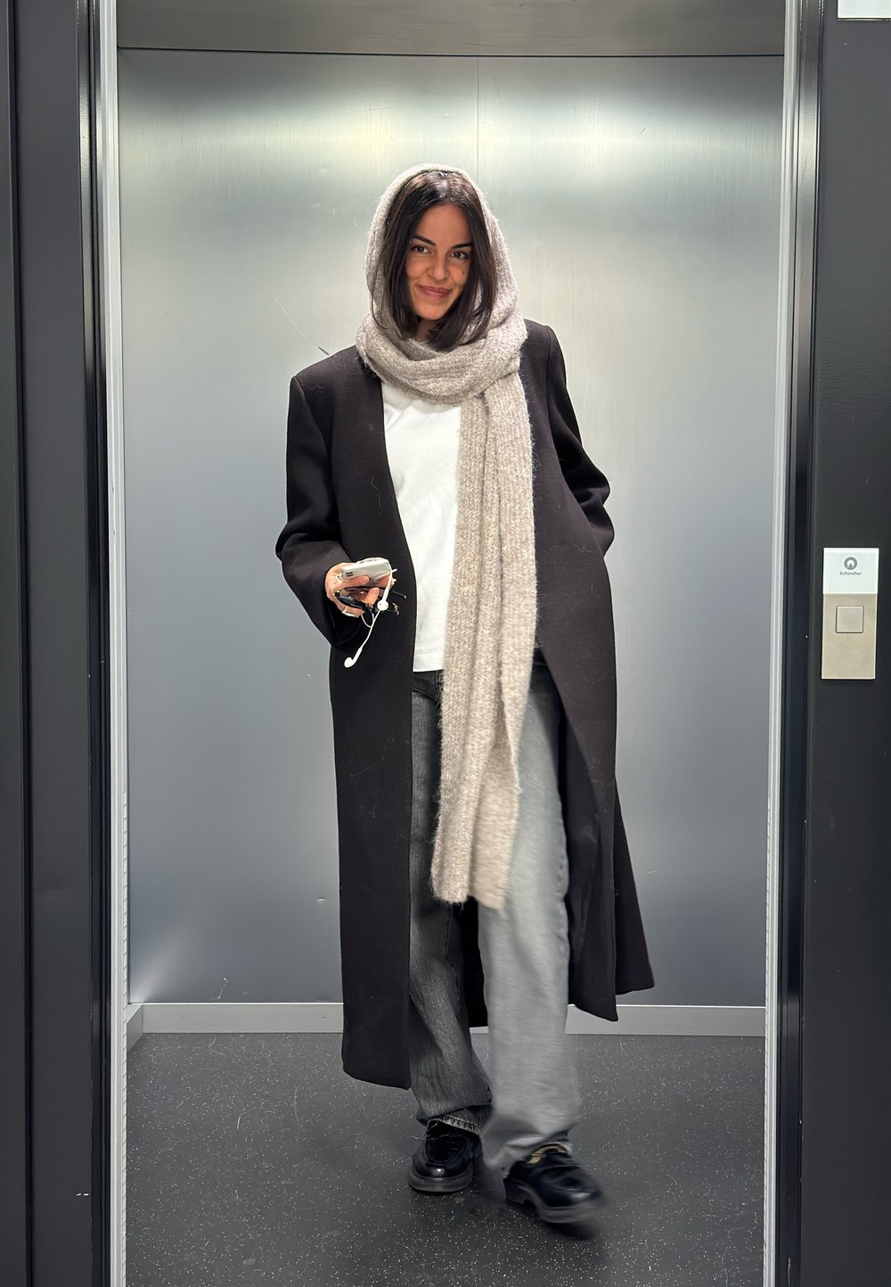 Kvinne som går ut av heisen iført lang svart frakk og grått skjerf, holder en telefon med hodetelefoner i hånden.