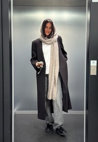 Kvinne som går ut av heisen iført lang svart frakk og grått skjerf, holder en telefon med hodetelefoner i hånden.