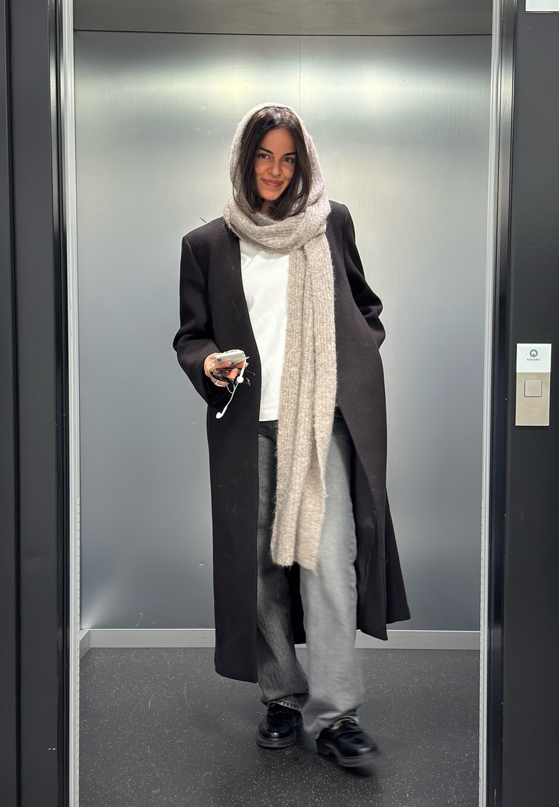 Vrouw die uit de lift stapt, gekleed in een lange zwarte jas en een grijze sjaal, met een telefoon met oordopjes in de hand.