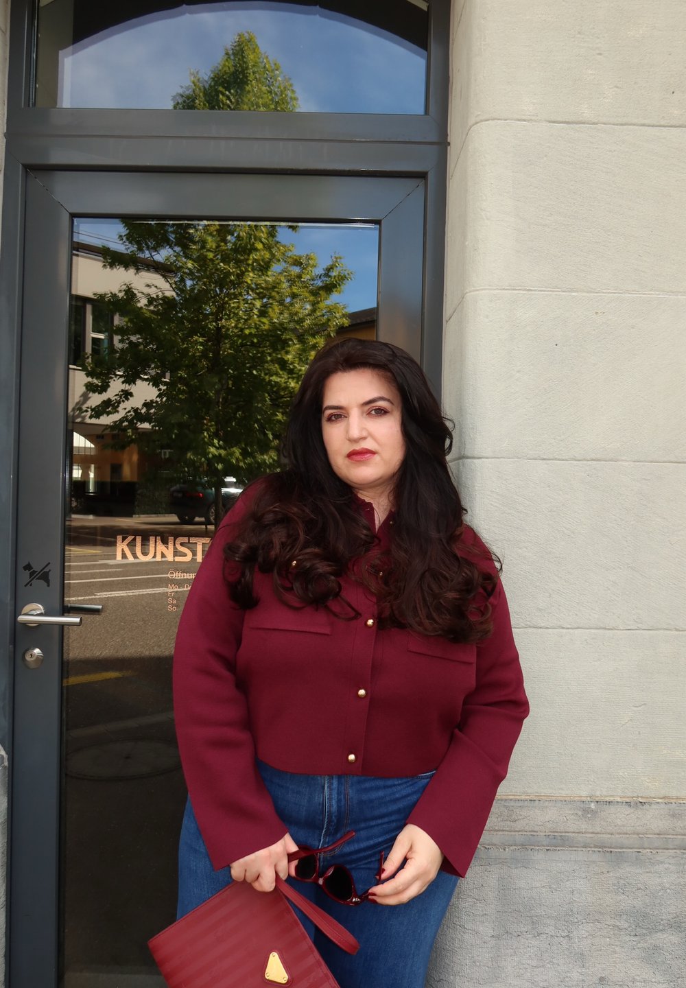 Kobieta z długimi ciemnymi włosami w maroonowej kurtce i niebieskich dżinsach, trzymająca pasującą kopertówkę i okulary przeciwsłoneczne, stojąca przy szklanych drzwiach z odbiciem drzewa.