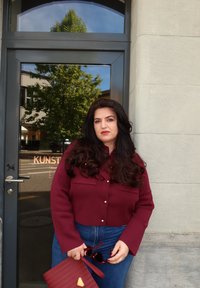 Kobieta z długimi ciemnymi włosami w maroonowej kurtce i niebieskich dżinsach, trzymająca pasującą kopertówkę i okulary przeciwsłoneczne, stojąca przy szklanych drzwiach z odbiciem drzewa.