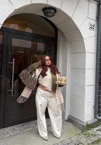 Beige neulottu villapaita, oliivinvihreä takki, kermaiset leveälahkeiset housut, ruudullinen huivi, kantaa kahta kahvikupillista tarjottimella, harmaan käsilaukun kanssa.