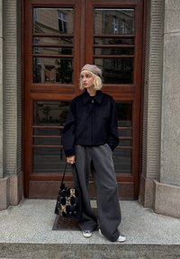 Svart zip-up jacka, vida grå byxor, vävd beige keps och mönstrad svart tote-väska med guldfinish. Släta texturer; neutral färgpalett.