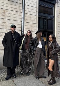 Quatre personnes se tiennent contre un mur texturé. Elles portent différents vêtements d'extérieur : un long manteau noir, des vestes en cuir et un imperméable en cuir marron. Parmi les accessoires notables, on trouve des lunettes de soleil et un béret.