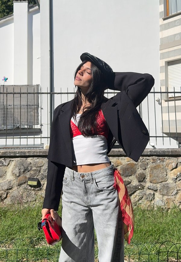 Jeune femme en jean à jambes larges, veste noire courte, top rouge et casquette noire, debout dehors tenant un sac rouge, les yeux fermés au soleil.