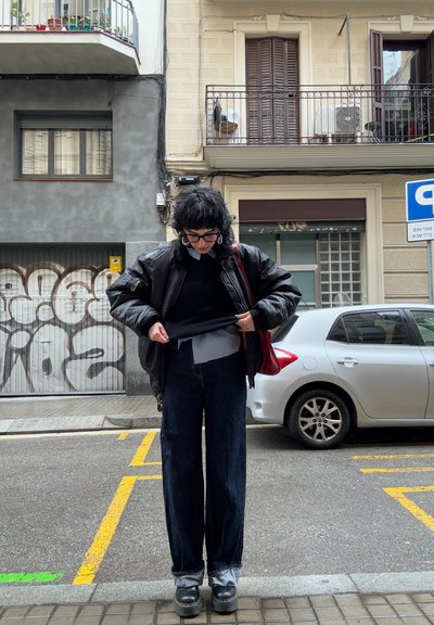 Chaqueta de cuero negra y gruesa combinada con un suéter negro y una camisa de color azul claro. Pantalones vaqueros oscuros y holgados, junto con zapatos negros gruesos, en una calle de la ciudad.