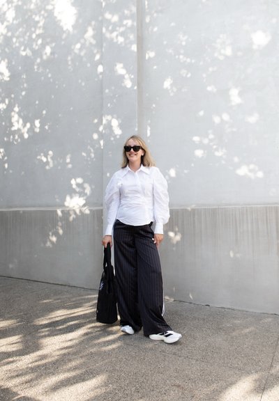 Camisa blanca de manga abullonada, pantalones anchos de rayas negras, zapatillas blancas y un bolso tote negro. El fondo presenta sombras suaves en una pared.