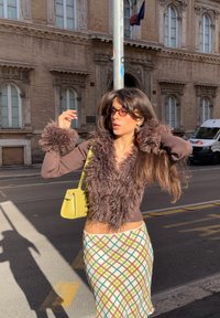 Brauner, flauschiger, kurz geschnittener Cardigan mit langen �Ärmeln, gemusterter beiger und grüner Rock und eine kleine gelbe Handtasche. Sonnenbrille komplettiert den Look.