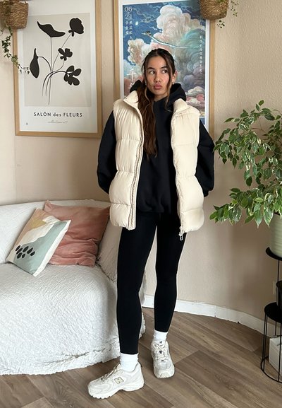 Chaleco blanco acolchado, sudadera negra, leggins negros y zapatillas ligeras. El fondo presenta plantas de interior y obras de arte enmarcadas.