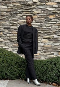 Zwarte oversized blazer over een zwart top en broek, met zilveren enkellaarzen. De achtergrond toont een stenen muur en groene bladeren.