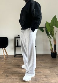 Fekete bomberdzseki cipzáros zsebbel, szürke melegítőnadrág és fehér sportcipő. A háttérben növényekkel és minimalista bútorokkal berendezett szoba.