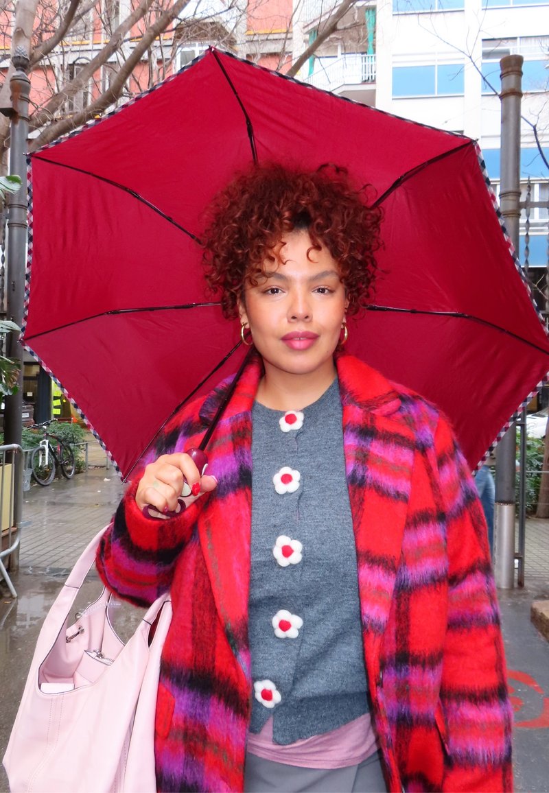 Femme aux cheveux bouclés portant un manteau à carreaux rouges et violets et un pull gris avec des boutons floraux, tenant un parapluie rouge dans une rue de la ville.