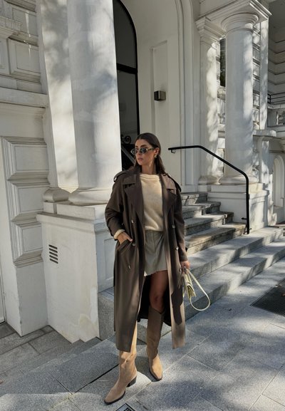 Abrigo largo marrón con botones de doble botonadura, suéter crema, pantalones cortos beige, botas altas beige y gafas de sol. Entorno urbano con escaleras.