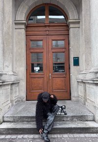 Personne en manteau de fourrure noire et chapeau assise sur des marches en pierre, ajustant des bottes noires à talons hauts, avec des portes doubles en bois et une façade de bâtiment en pierre grise en arrière-plan.