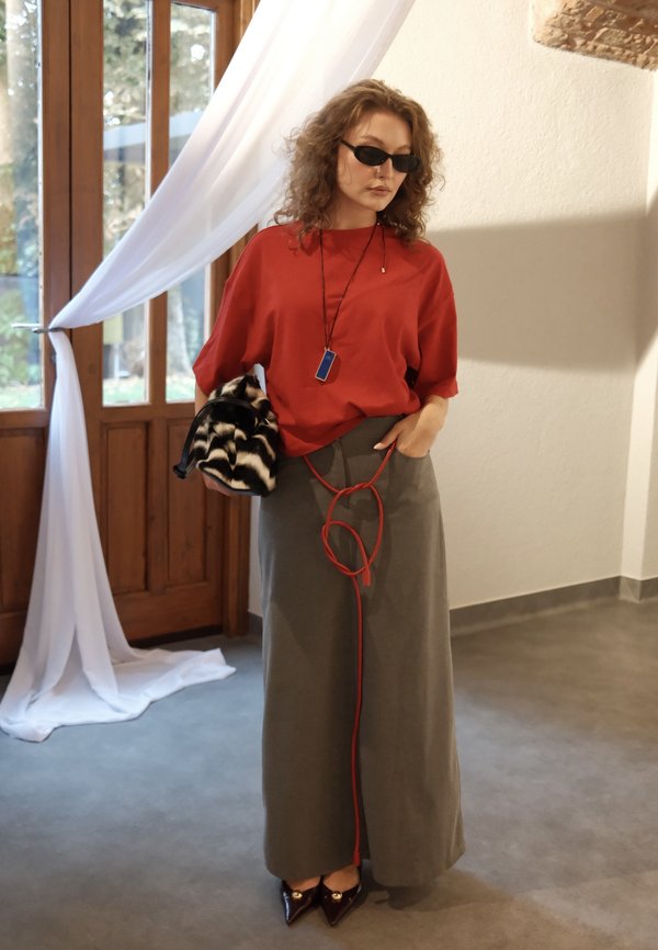 Donna con top rosso, gonna lunga grigia con cintura corda rossa, occhiali da sole neri, che tiene una pochette nero e bianco con motivo all'interno vicino a porte di legno.