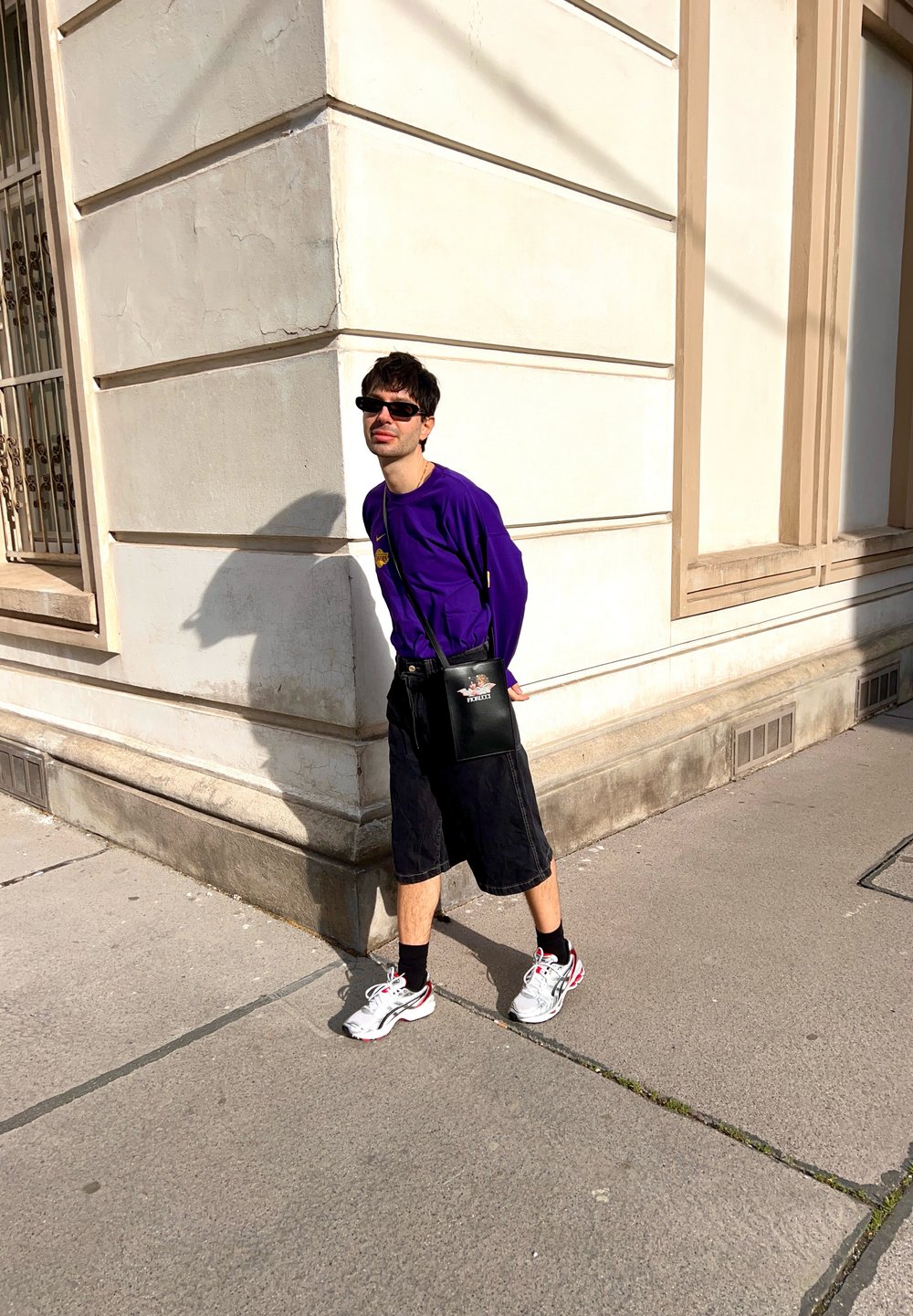 Una persona con una maglietta a maniche lunghe viola, pantaloni corti neri, borsa nera e scarpe da ginnastica grigie si trova all'angolo di un edificio, mentre la luce del sole proietta un'ombra.