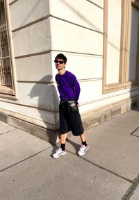 Una persona con una maglietta a maniche lunghe viola, pantaloni corti neri, borsa nera e scarpe da ginnastica grigie si trova all'angolo di un edificio, mentre la luce del sole proietta un'ombra.