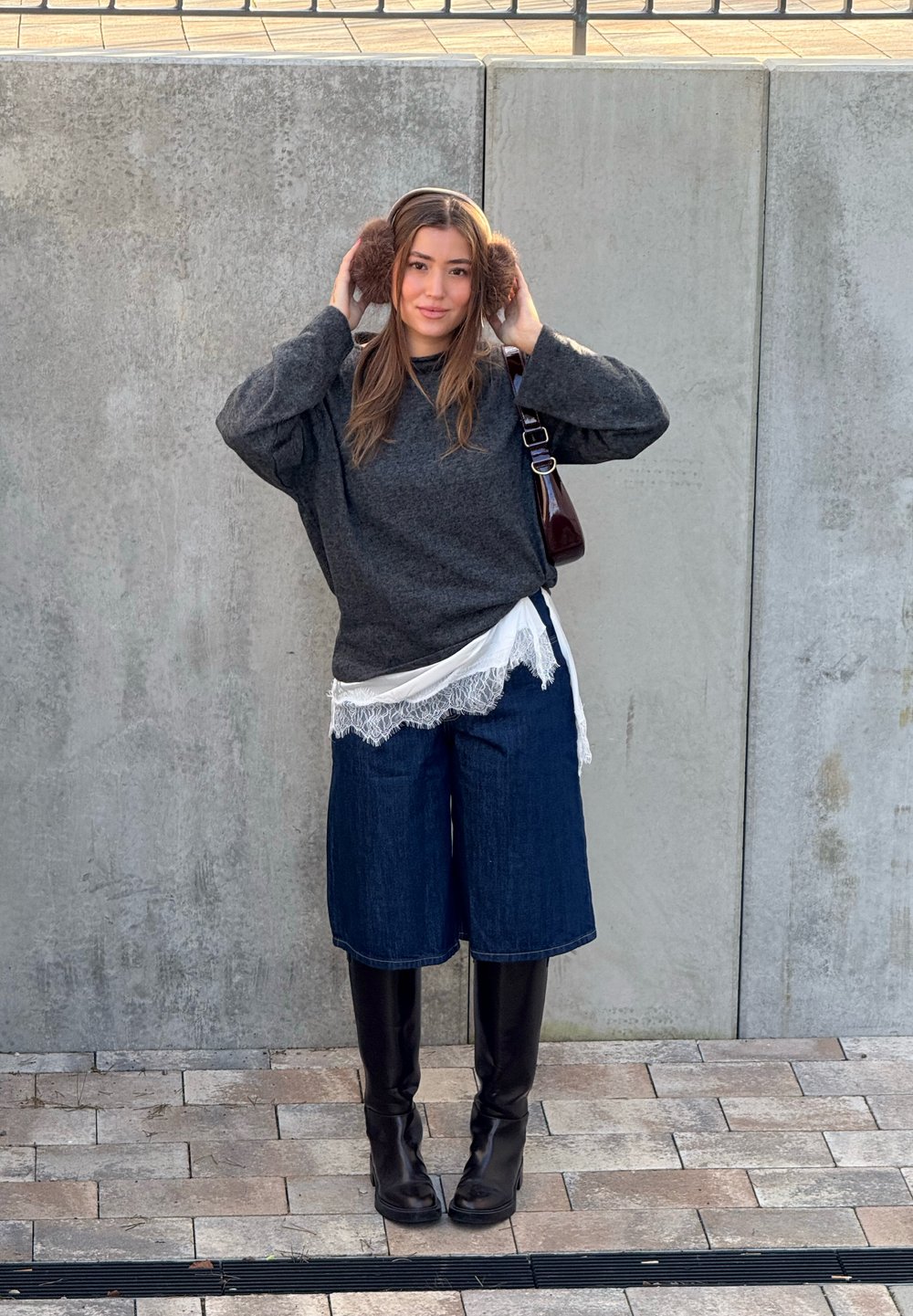 Femme ajuste ses cache-oreilles marron, portant un pull gris foncé, un haut en dentelle blanche, un short en jean, des bottes noires montant jusqu'aux genoux, debout sur un sol carrelé à côté d'un mur en béton.