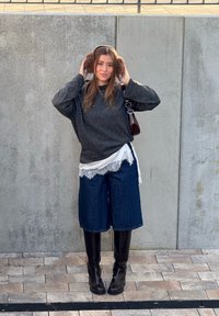 Femme ajuste ses cache-oreilles marron, portant un pull gris foncé, un haut en dentelle blanche, un short en jean, des bottes noires montant jusqu'aux genoux, debout sur un sol carrelé à côté d'un mur en béton.