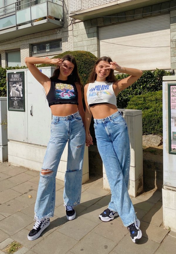 Dos personas están juntas, vistiendo tops cortos y jeans rotos de un azul claro. Llevan zapatillas en blanco y negro y posan haciendo signos de paz.