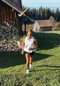 Femeie alergând în sus pe o potecă erbacee, lângă lemne de foc stivuite și cabane din lemn, cu copaci și dealuri în fundal.