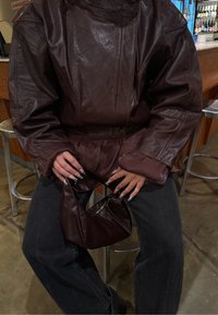 Ruskea nahkainen ylisuurikokoinen takki, jossa on vyötäröllä kiristys, yhdistetty tummansinisiin farkkuihin. Henkilöllä on pieni ruskea nahkainen laukku ja pitkät, koristellut kynnet.