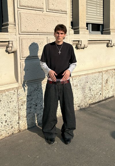 Camisa negra de manga corta sobre una camisa gris de manga larga, pantalones negros de rayas oversize, cinturón rojo con hebilla metálica y zapatos negros.