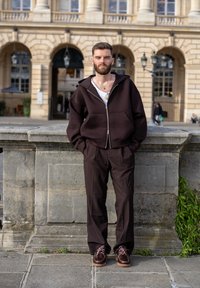 Dunkelbrauner Reißverschluss-Hoodie mit großen Taschen und Kapuze, kombiniert mit passenden Hosen und braunen Schuhen mit pinken Schnürsenkeln auf einem steinernen Hintergrund.