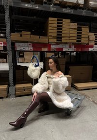 Crème faux fur jas met brede mouwen, gedragen over een witte jurk. Bruine haklaarzen en een witte faux fur handtas met blauwe accenten op een winkelwagentje.