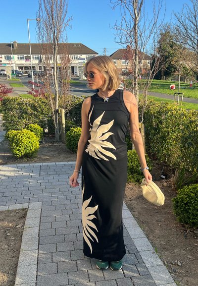 Vestido maxi negro con patrón de hojas beige; diseño sin mangas y ajuste ceñido. Accesorios con un pequeño bolso de paja y zapatos verdes.