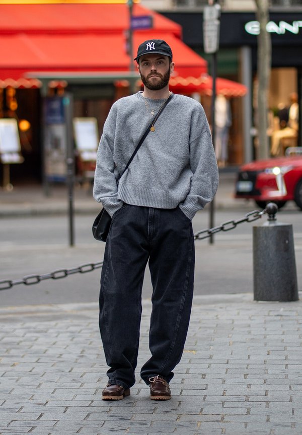 Homme portant un pull gris, un pantalon noir ample, des chaussures marron foncé et une casquette noire, debout dans une rue de la ville avec un auvent rouge en arrière-plan.