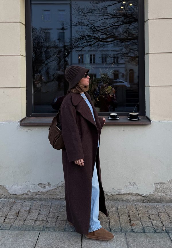 Femme en manteau long marron et chapeau debout près d'une fenêtre de café avec deux tasses de café sur le rebord, la rue de la ville se reflète dans la vitre.