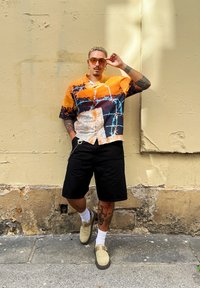 Κοντομάνικο πουκάμισο με πορτοκαλί, μαύρο και λευκό αφηρημένο σχέδιο, συνδυασμένο με μαύρες βερμούδες, μπεζ παντόφλες και λευκές κάλτσες.