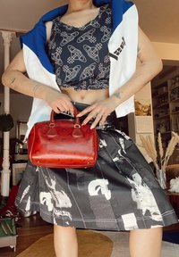 Červená kožená kabelka s zaoblenými ušami, v ktorej sa nachádza čierna sukňa s grafickým vzorom a modro-biela crop top s uzlom.