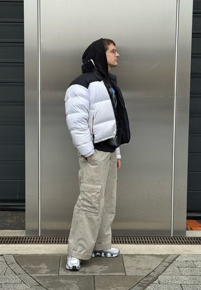 Chaqueta acolchada con paneles en blanco y negro, sudadera con capucha gris debajo, pantalones cargo beige y zapatillas plateadas metálicas. De pie frente a un fondo metálico.