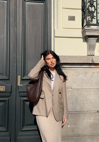 Kremo spalvos pritaikytas švarkas ant baltos palaidinės, derinamas su figūrą ap_trialsiančia kremo spalvos sijonu; laiko rudą pintą rankinę prieš tamsias duris.