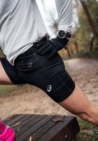 Fekete futó rövidnadrágot, fehér hosszú ujjú inget és kesztyűt viselő személy nyújtja a lábát egy fa padon egy szabadtéri, őszi környezetben.