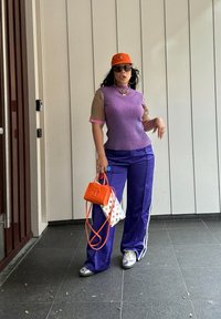 Fialový pletený top s vertikálními pruhy a krátkými rukávy, fialové saténové kalhoty s bílými bočními pruhy, oranžová čepice a tašky v oranžové a bílé barvě s puntíky.