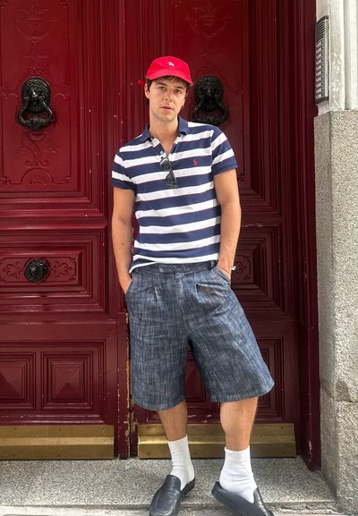 Camisa polo a rayas azul y blanca, pantalones cortos oscuros, gorra roja, zapatos negros y calcetines blancos, frente a una puerta roja profunda con aldabas en forma de cabeza de león.