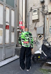 Άνδρας που φοράει πράσινο και μαύρο μπουφάν με σχέδιο κρανίου στέκεται απέναντι από έναν τοίχο με ηλεκτρικά κουτιά και πινακίδες προειδοποίησης δίπλα σε μια λευκή σκούτερ.