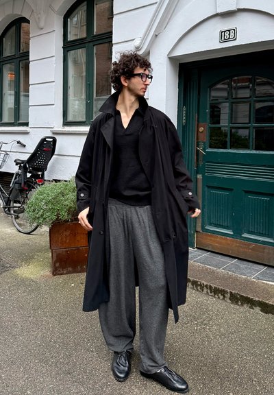 Abrigo negro tipo gabardina sobre un suéter negro, pantalones de pierna ancha grises y zapatos negros. El conjunto presenta un estilo casual y relajado. Puerta verde al fondo.