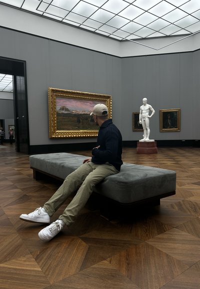 Banco tapizado en gris con patas de madera oscura, colocado sobre un suelo de madera, junto a obras de arte enmarcadas y una escultura blanca en un ambiente de galería.