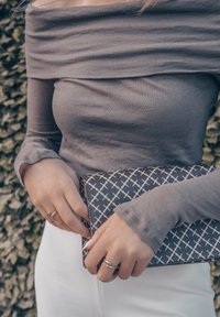 Γκρι τοπ με ανοιχτούς ώμους και μακριά μανίκια, κρατώντας μια τσάντα με μαύρο και άσπρο σχέδιο, σε συνδυασμό με λευκά παντελόνια. Ορατό υφασμάτινο σχέδιο.