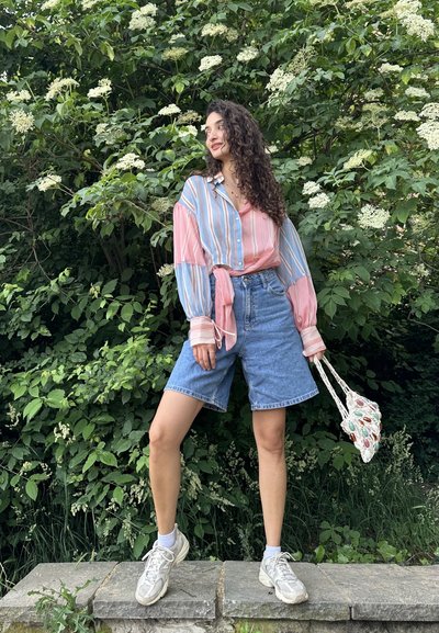 Mujer con cabello largo y rizado está de pie sobre un saliente de piedra frente a arbustos verdes con hojas y flores blancas, lleva una camisa de rayas y pantalones cortos de mezclilla.